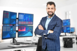 Smiling male Sydney Cloud IT Services expert standing in front of desk with four monitors - Contact Sydney Managed IT Services today