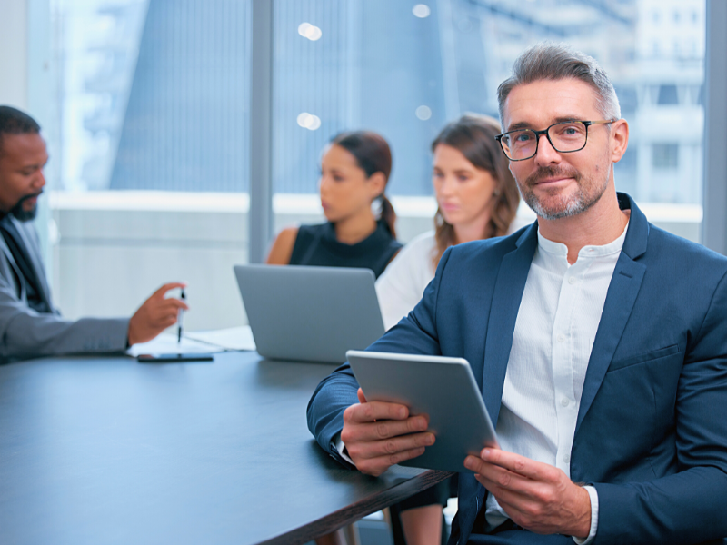 Sydney Manage IT Best IT Service In Australia Businessman holding a tablet looking in front of a camera, colleagues having a meeting in the background