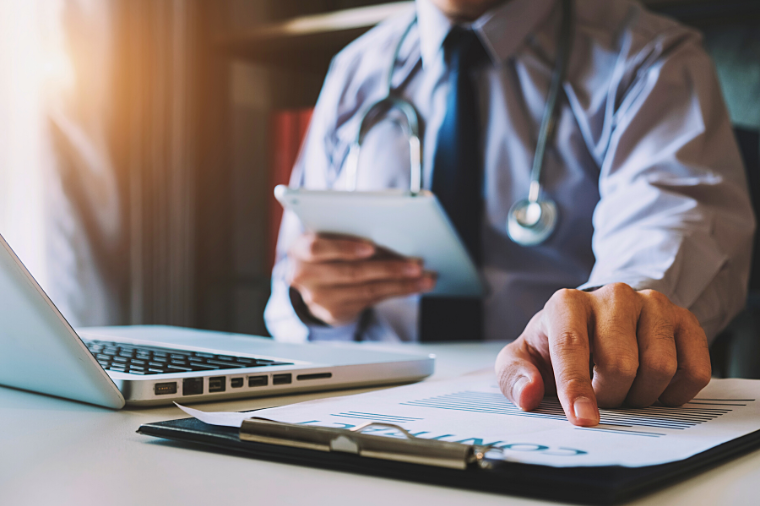 Contact Sydney Managed IT Experts In Medical IT Cropped image of a doctor checking a patient's medical information on a clipboard and tablet, laptop can also be seen on the table
